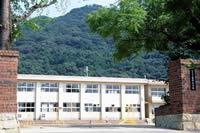 山の麓に立つ白壁で2階建ての校舎を、レンガ造りの校門外から撮影した龍野小学校の写真