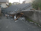 屋根のみを残して潰れた家と回りの塀も崩れてしまっている写真