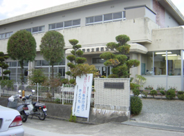 2階建ての白の外観で、周りには数本の植木が植えられた公民館入口付近の外観写真