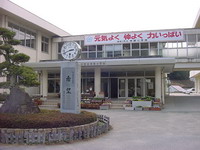 大きな石や時計が設置された円形の花壇の奥にある玄関前の階段に花が植えられたプランターが並び、2階建ての校舎の外側に「元気よく 仲よく 力いっぱい」と書かれた横幕が貼られた神部小学校の写真