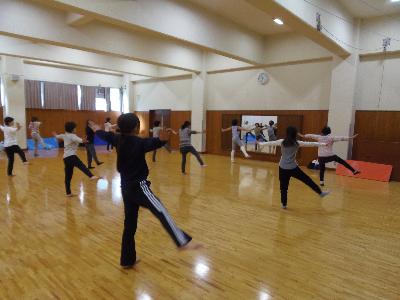 14人程の参加者が前にいる講師のポーズを真似して、両手を広げ、右足を上げ左足でバランスをとっているバレトンの写真