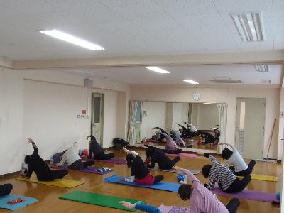 ヨガマットを並べその上で右足を曲げ、右手で左足のつま先を触ろうする参加者と1人の方に講師がアドバイスをしていピラティスの写真