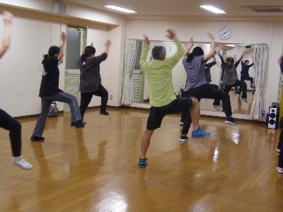 鏡の前に指導者が立ちダンスの見本を見せ、参加者が立って両手を上にあげ、右足を引き上げてダンスを行っているリズムダンスの写真