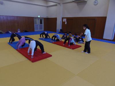 マットの上でブリッジをし、その前で指導者がアドバイスをしている小学生体操の写真