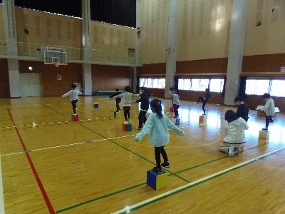 体育館の前方で幼児と向かい合わせになり講師が見本を見せているのを幼児が3列程に並び両足がのるくらいの正方形の箱の上で両手を広げ左足だけで立っている幼児体操の写真