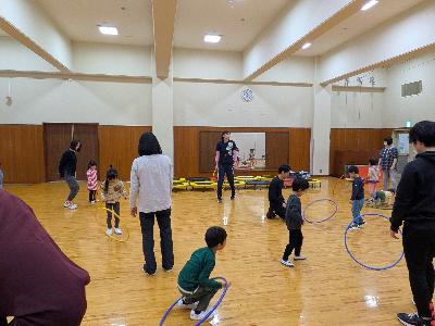 参加者がフラフープを跳んだり回したりしている前で講師が指導しているキッズスポーツリズムの写真