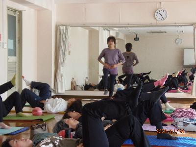 ヨガマットを並べその上で左足は足裏を地面につけたまま90度に曲げ、右足はふくらはぎと床を平行に保つポーズをする参加者と1人の方に講師がアドバイスをしていピラティスの写真