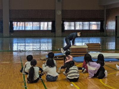 講師が跳び箱を跳ぶ見本をする前で、小学生が跳び方を見学している小学生体操の写真