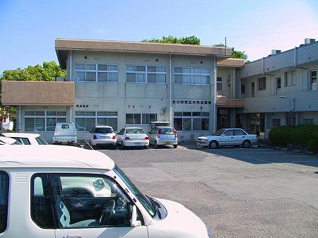 二階建ての小宅公民館の建物と、その建物の前にある複数台の車がとめられた駐車場を写した写真