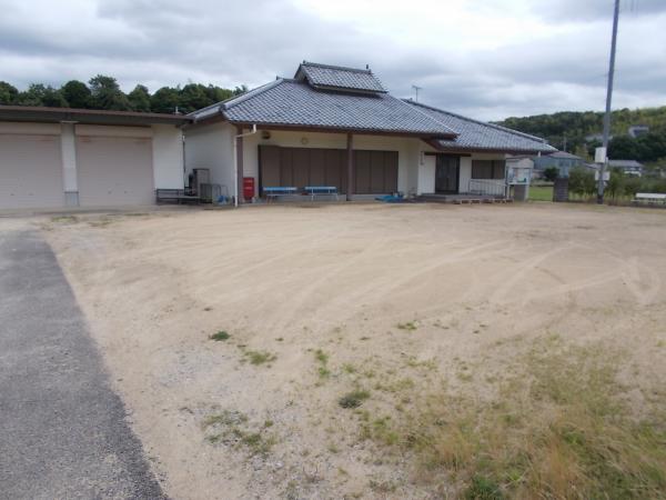 瓦屋根と白い外壁の一階建ての小畑農業構造改善センターの建物と、その建物前にある土の広い駐車スペースの写真