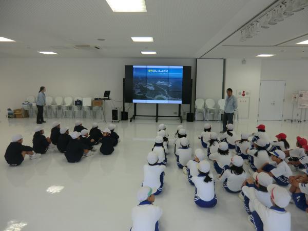 白い内装の部屋で前方に設置されているモニターに動画を流しながら説明している職員の方と、床に並んで座っている児童たちの写真