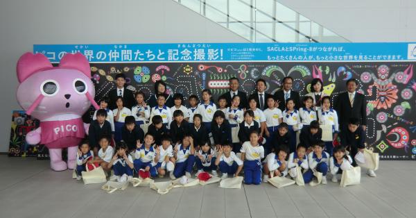 記念撮影用の長方形の看板の前で、ピンク色の猫の着ぐるみと一緒に写真を撮っている、整列した児童たちとスーツを着た大人の方々の写真
