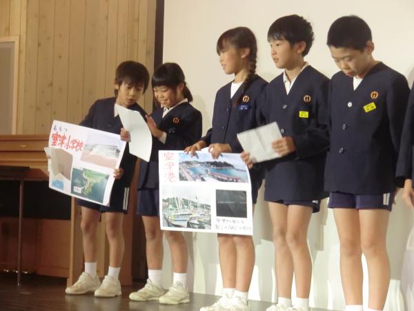 紺色の制服を着た児童たちが、ステージの上で手作りの資料を手に持って発表をしている写真