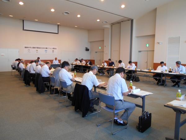 タイル模様のカーペットの敷かれた広い会議室で、四角く机を並べて会議をしている職員の方々の写真