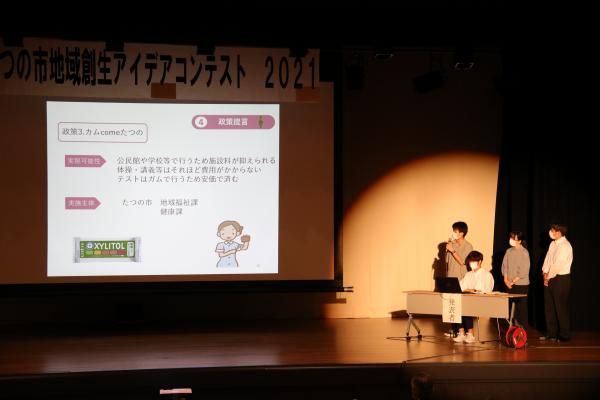 たつの市地域創生アイデアコンテスト2021と書かれた幕の張られているステージの上で、スクリーンに映像を映しながらその脇で説明をしている人たちの写真