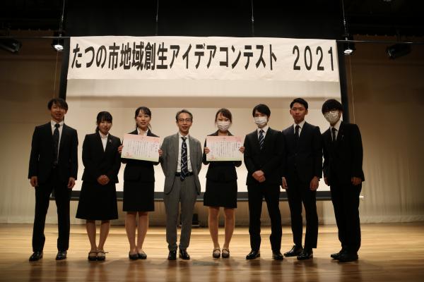 たつの市地域創生アイデアコンテスト2021と書かれた幕の張られているステージの上で、グレーのスーツを着た方の両脇に賞状を持った「ウェルネス創業チーム」、「エリアリノベーションチーム」が並んでいる写真