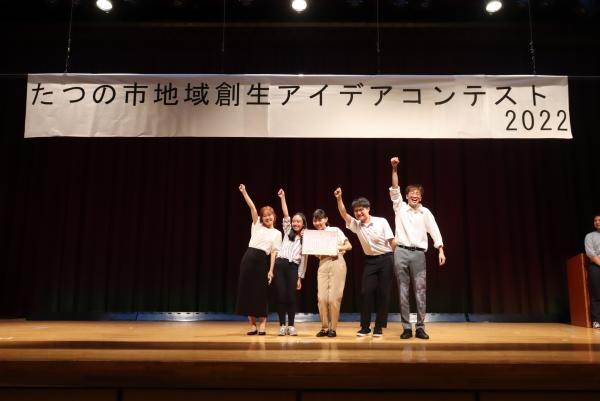 優秀賞を受賞した大阪大学赤井研究室大橋班の皆さんが、舞台の上で賞状を持ってぎゅっと握りしめた手を上にあげるポーズをとっている写真