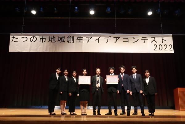 最優秀賞を受賞したチームの皆さんと審査員特別賞を受賞したチームの皆さんが、それぞれ賞状を持ち一緒に横一列に並んで舞台上に立っている写真