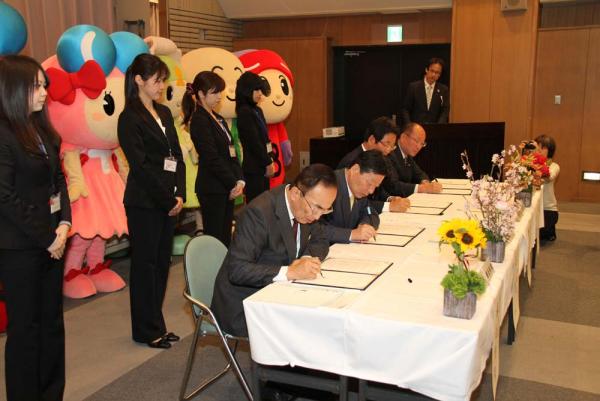 白いテーブルクロスが敷かれた長机で、黒いスーツを着た秘書の方たちが後ろに控えている、協定書にサインをしているスーツを着た男性たちの写真