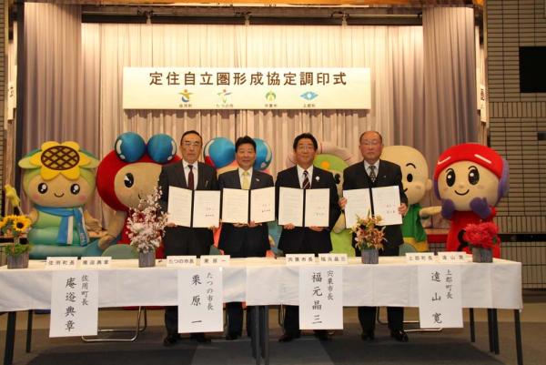 定住自立圏形成協定調印式と書かれた幕の張られたステージの前で、協定書を両手で持って並んでいるスーツを着た4人の男性の写真