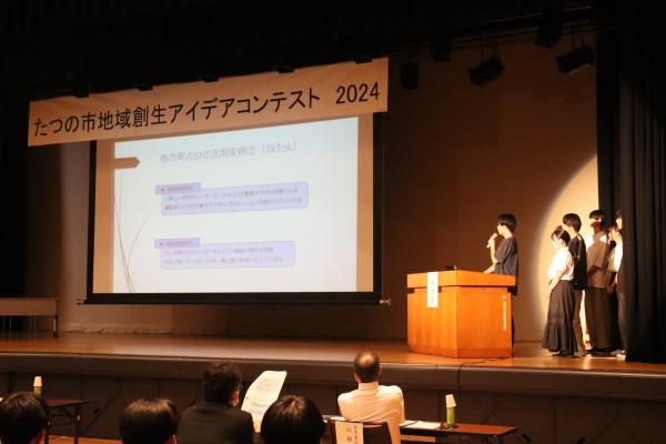 京都産業大学SNS発信チームの学生さんたちが、舞台の上に立ち大きなスクリーンに映される資料の方を向き話をしている様子の写真