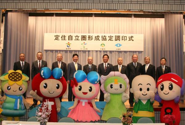 定住自立圏形成協定調印式と書かれた幕の張られたステージの前で、着ぐるみのマスコットキャラクターとスーツを着た男性たちが並んでいる写真