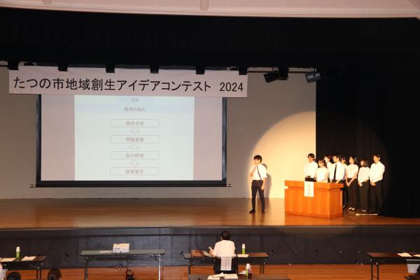 大阪大学赤井ゼミの学生さんたちが、舞台の上に立ち大きなスクリーンに映される資料とともに発表を行っている写真