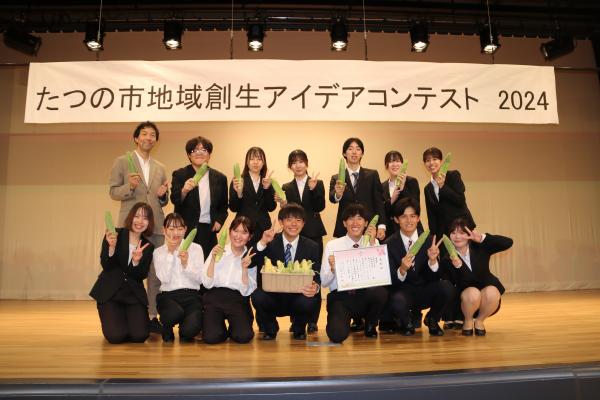 優秀賞を受賞した兵庫県立大学Bチームの皆さんが、舞台の上で賞状や薄緑色の野菜のようなものを持って記念写真を撮っている写真